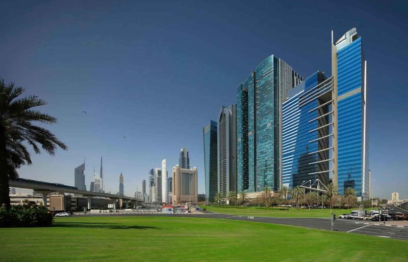 Modern blue glass skyscrapers line a grassy park area with palm trees in a city skyline. In the foreground, a road and vehicles frame the scene.