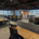 Open-plan office with long wooden desks, black ergonomic chairs, and partitions, large windows overlook a city skyline after daylight.
